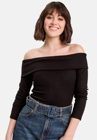 Giovane donna che sorride, indossando un top a spalle scoperte in nero a costine e jeans blu a vita alta con una cintura larga, su uno sfondo bianco uniforme.