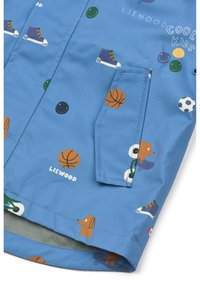 Sinine vettpidav jope, millel on värvikad sporditeemalised mustrid, sealhulgas korvpallid ja multifilmikoerad, ning küljetasku detail.