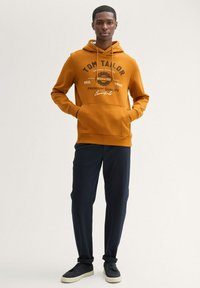Hanorac portocaliu cu glugă și buzunar frontal, având text grafic alb și negru. Asortat cu pantaloni tapered de un albastru închis și teniși negri.