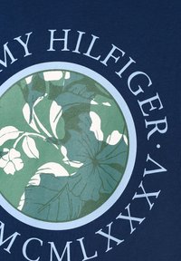 Námořnické tričko s kruhovým zeleným květinovým grafikem, s textem "TOMMY HILFIGER" v bílé barvě nahoře a římskými číslicemi níže.