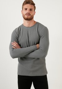 Siva dolgi rokav riban t-shirt z okroglim izrezom in prilegajočimi rokavi, v kombinaciji s črnimi hlačami. Tekstura je rahlo dvignjena.