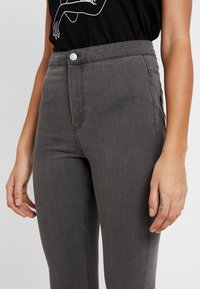 Jeans de tiro alto grises en tela de mezclilla, con cierre frontal de botón, bolsillos laterales y detalles de costura visibles.