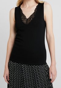 Černý top bez rukávů s výstřihem do V, zdobený černou krajkou. Top je přiléhavý, vyrobený z hladké látky, v kombinaci s sukní s puntíky.