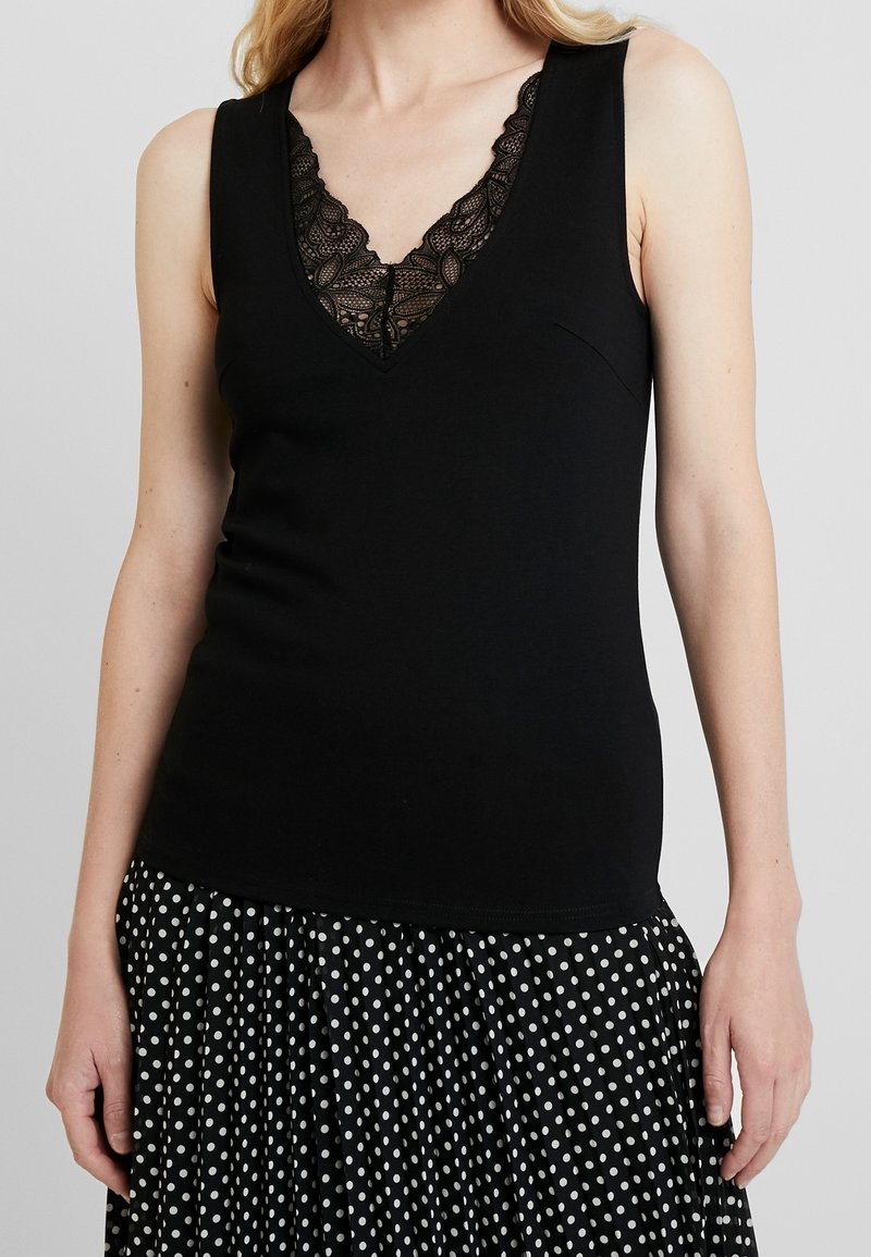 Černý top bez rukávů s výstřihem do V, zdobený černou krajkou. Top je přiléhavý, vyrobený z hladké látky, v kombinaci s sukní s puntíky.