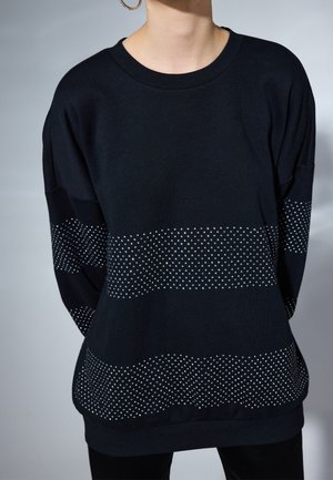 Osoba ubrana w czarny sweter z trzema poziomymi pasami drobnych białych kropek na wysokości klatki piersiowej, rękawów i dołu.