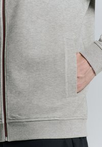 Lichtgrijze zip-up jas van zachte stof; heeft een handzak en ribgebreide manchetten. De binnenkant van de rits is rood.