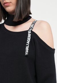 Black knit sweater with a high collar and one cold-shoulder design. Features a prominent black and white logo strap. Soft texture.