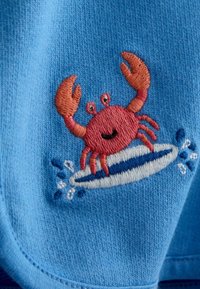 Rød broderet krabbe med løftede kløer, der surfer på et stribet blå og hvidt surfbræt med vandsprøjt på lyseblåt stof.