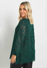 Grön spetsblus med långa ärmar, blommönster, fjärilskant nertill och transparent design, tillsammans med svarta byxor.