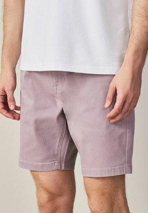 Férfi világoslila rövidnadrágban és fehér pólóban, laza tartással, karjai oldalt, semleges háttér előtt áll.