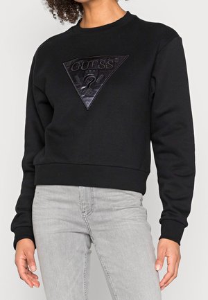 Persoon draagt een zwarte Guess-trui met een reliëf driehoekig logo en lichtgrijze spijkerbroek, staand met ontspannen armen.