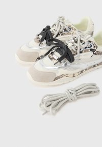 Αθλητικά παπούτσια με μείγμα υλικών που περιλαμβάνουν μπεζ σουέντ, μεταλλικό ασημί και λεπτομέρειες με φίδι. Μαύρα και γκρι κορδόνια, υφασμάτινη λεπτομέρεια με υφή δικτυώματος.