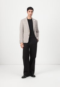 Hellgrau, strukturiertes Blazer mit braunen Knöpfen, kombiniert mit einem schwarzen Hemd und schwarzen, weit geschnittenen Hosen. Schwarze Schuhe runden das Outfit ab.