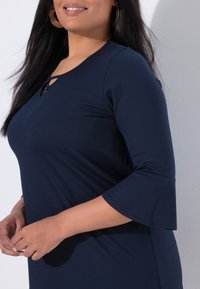 Kobieta o długich ciemnych włosach, ubrana w granatową bluzkę z rozszerzanymi rękawami długości do łokcia oraz małym wycięciem przy dekolcie.