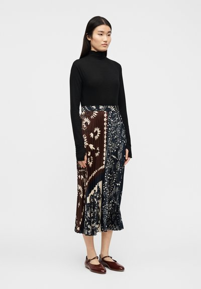 Γυναίκα με μαύρο τοπ μακρυμάνικο και πλισέ μίντι φούστα με  καφέ και ναυτικό μπλε σχεδιασμό παϊέτας, φοράει σκούρα καφέ παπούτσια Mary Jane.