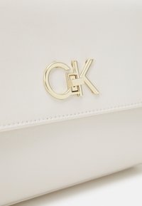 Bílá taška z umělé kůže s logo "CK" v zlaté barvě a hladkou, matnou texturou se subtilním prošitím podél okrajů.