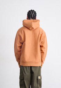 Kobberfarvet hoodie med hætte, lavet af blødt stof, med en løs pasform og ribbede manchetter. Kombineret med olivengrønne cargo bukser.