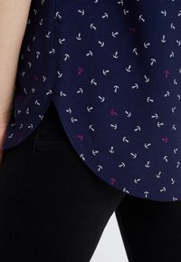 Mörkblå topp med ett upprepande ankarmönster i vitt, rosa och lila. T-shirten har en rundad nederkant och en mjuk tygtextur.