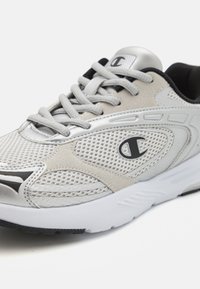 Lehká sportovní obuv kombinující šedou síťovinu a stříbrný syntetický materiál, s černými detaily a gumovou podrážkou.