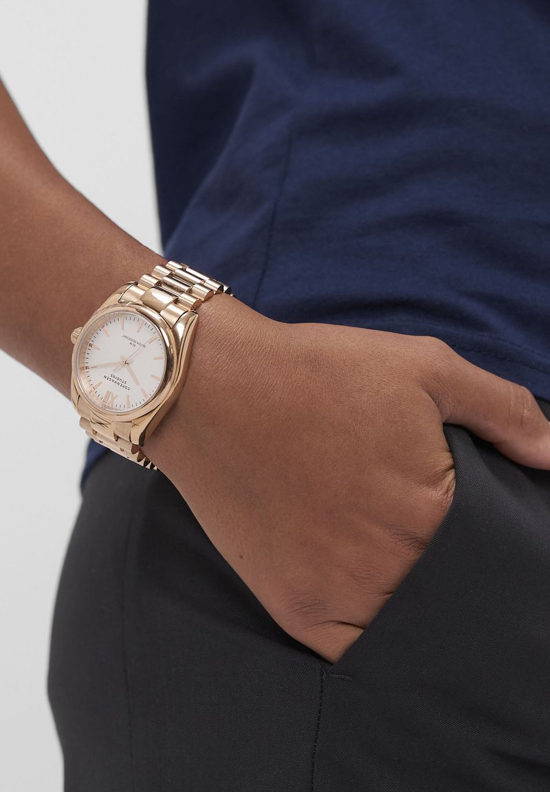 Braunhäutige Hand in der Tasche einer schwarzen Hose, die eine goldene Metallband-Armbanduhr mit weißem Zifferblatt und blauer Oberseite trägt.