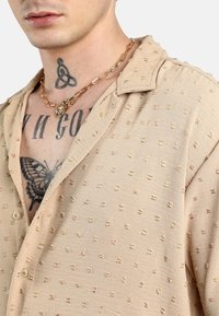 Camisa beige con superficie texturizada, pequeños patrones en relieve y un collar. Presenta botones y un escote abierto, acompañada de un collar de cadena dorada.