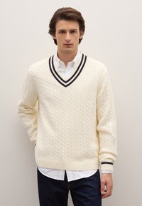 Crèmekleurige kabelgebreide trui met een V-hals en donkere gestreepte accenten, gedragen over een witte blouse met kraag en gecombineerd met donkere jeans.