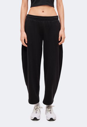 Femme portant un pantalon de survêtement noir ample, des baskets blanches et un top court noir, montrée de la taille jusqu'aux pieds sur un fond uni.