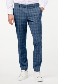 Man som bär blå rutig slim-fit-byxor, vit skjorta och vita sneakers, står med en hand i fickan.