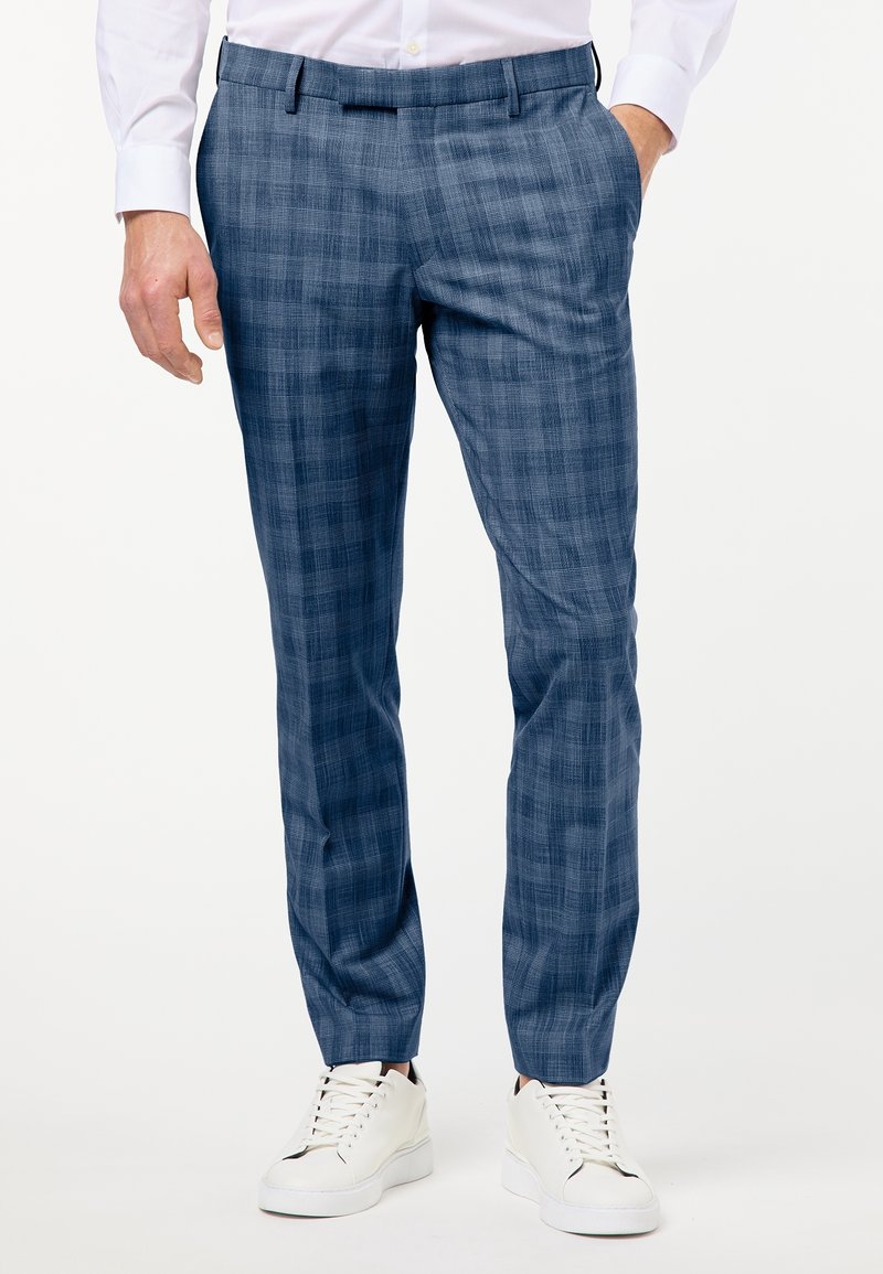 Man som bär blå rutig slim-fit-byxor, vit skjorta och vita sneakers, står med en hand i fickan.