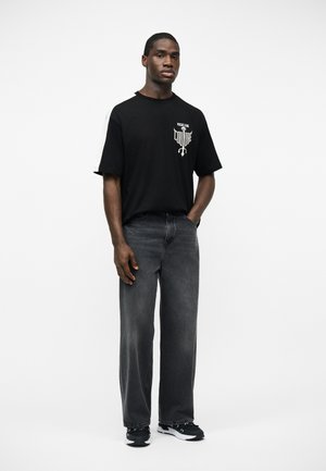 Άνδρας με το χέρι στην τσέπη, φοράει μαύρο t-shirt Versace Jeans Couture, ξεθωριασμένο μαύρο παντελόνι φαρδιάς γραμμής και μαύρα αθλητικά παπούτσια, μπροστά σε λευκό φόντο.