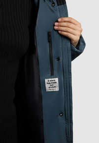 Blauwe pufferjas met een zachte textuur, voorzien van een zwart geribbeld voering, zijzak met rits, en een label met de tekst "Een warme omhelzing van stof."