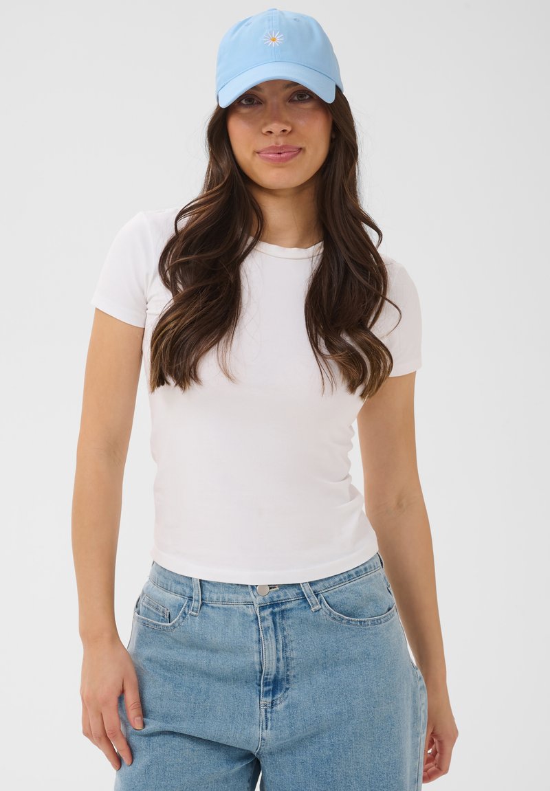 Jeune femme aux longs cheveux ondulés portant un T-shirt blanc ajusté, un jean bleu clair et une casquette de baseball bleu clair avec une petite marguerite.