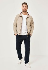 Light beige bomber jacket with a red and navy collar, zip pockets, worn over a white shirt, paired with dark navy pants and white sneakers.