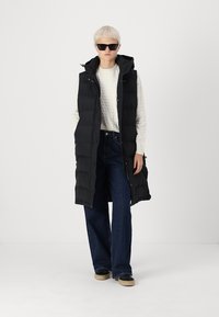 Pitkä musta puffatuliivi hupulla, jossa on nappikiinnitys, käytetty kevyen teksturoidun neuleen ja leveälahkeisten tummien farkkujen päällä.