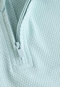 Tejido texturizado de color azul claro con un patrón de diamantes; presenta una cremallera gris visible y un diseño de cuello con cremallera.
