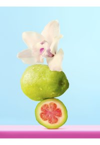 Guayaba verde con gotas de agua, coronada con una flor de orquídea blanca, equilibrada sobre una mitad de guayaba cortada, sobre un fondo azul pastel.