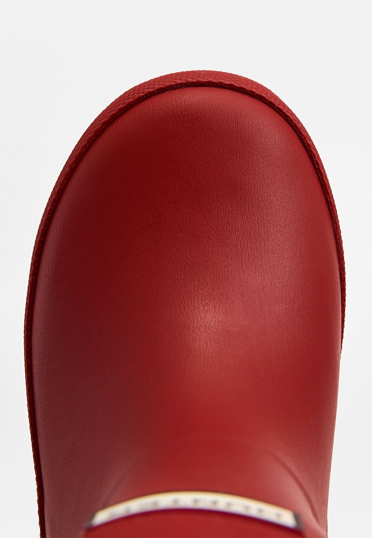 Botte en caoutchouc rouge avec une surface lisse et brillante, un bout arrondi et une semelle texturée. Comprend un petit accent près du talon.