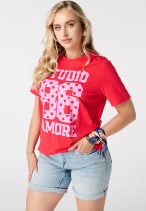 Femme aux longs cheveux blonds portant un t-shirt rouge avec le texte « "STUDIO 86 AMORE" » et un short en jean clair, posant une main dans la poche.