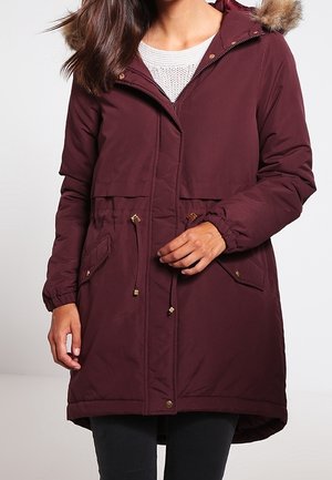 Femme portant un manteau d'hiver à capuche bordeaux avec cordons, poches à rabat et bordure en fausse fourrure sur la capuche.