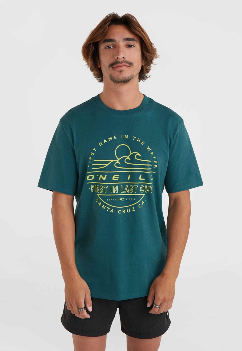 Teal bomulds t-shirt med gul grafisk tekst og bølgedesign. Korte ærmer, rund hals, afslappet pasform, sammen med sorte shorts.