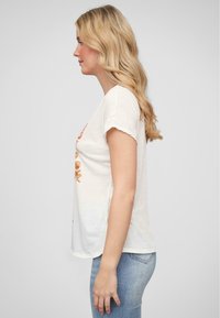 T-shirt en coton blanc avec un design graphique sur le devant, manches courtes et un ourlet légèrement arrondi, associé à un jean bleu clair.