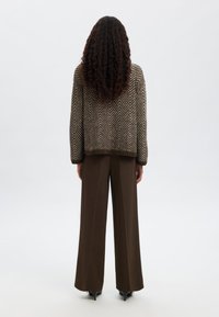 Rjav pulover iz pletenine z vzorcem ribje kosti in sproščenim krojem, kombiniran s širokimi rjavimi hlačami in črnimi čevlji z visokimi petami. Bele ozadje.