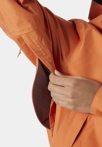 Chaqueta naranja hecha de una tela ligera y texturizada, con un forro interior de un tono más oscuro, que presenta bolsillos con cremallera y puños ajustables.
