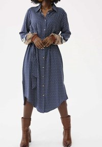 Femme portant une robe midi à boutons bleu marine à motifs avec des poignets à motifs contrastants et des bottines à talons marron, debout.