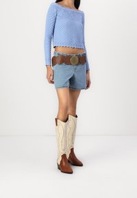 Blå stickad off-shoulder croptop, denimshorts, brunt läderbälte med stort spänne och beige cowboyboots med dekorativt broderi.