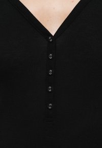 Top nero in maglia coste con scollo a V e patta frontale con bottoni metallici, creando un aspetto strutturato e aderente.