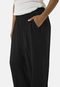 Pantalones negros, ligeros y de corte relajado, con una cinturilla ancha y bolsillos laterales. La tela tiene una textura sutil.