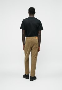 Pantaloni din corduroy maro cu un finisaj texturat, având buzunare spate și o etichetă din piele bej la talie, purtați cu un tricou negru.