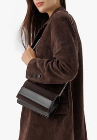 Borsa in suede marrone con tracolla in metallo argentato, presenta un coperchio ripiegato in una finitura scura a contrasto e dettagli di superficie testurizzati.