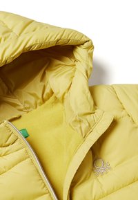 Žlutá prešívaná bunda s kapucí, která má měkkou fleecovou podšívku a malé vyšívané logo. Zapínání na zip a hladká struktura.